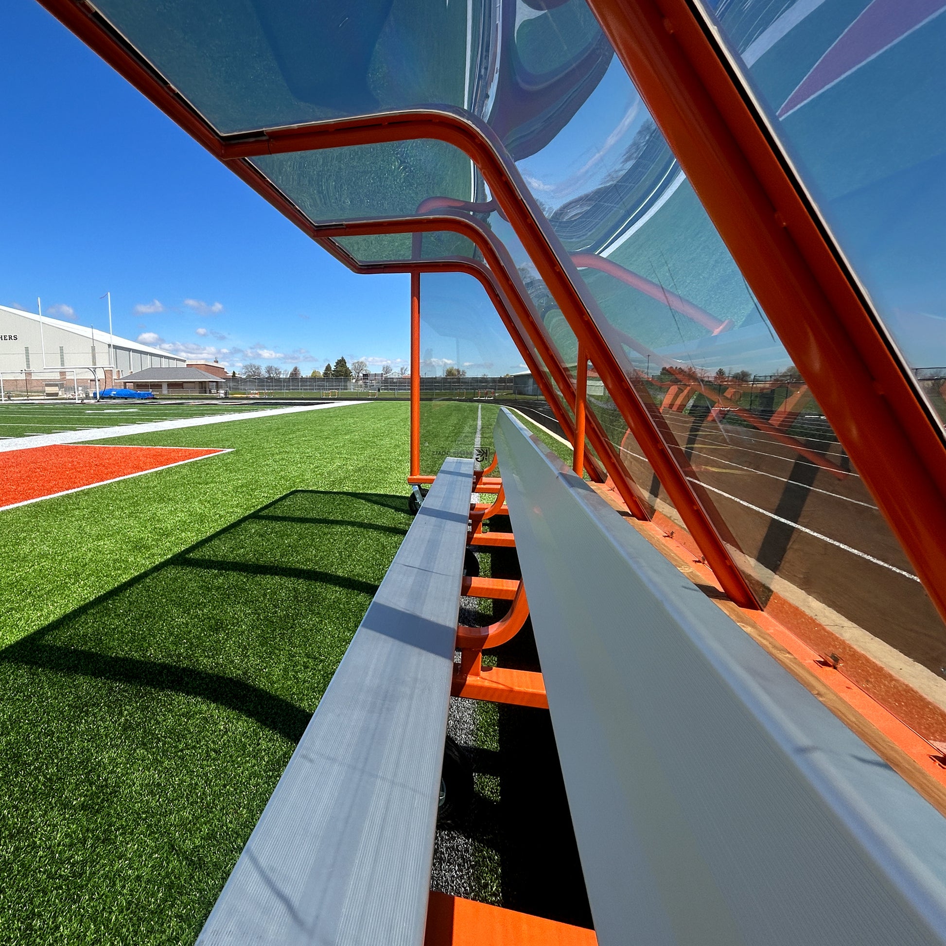 A photo of the 16 Traditional Heavy-Duty Team Shelter with a clear, orange-framed canopy beside an artificial turf field under a blue sky at an outdoor stadium.
