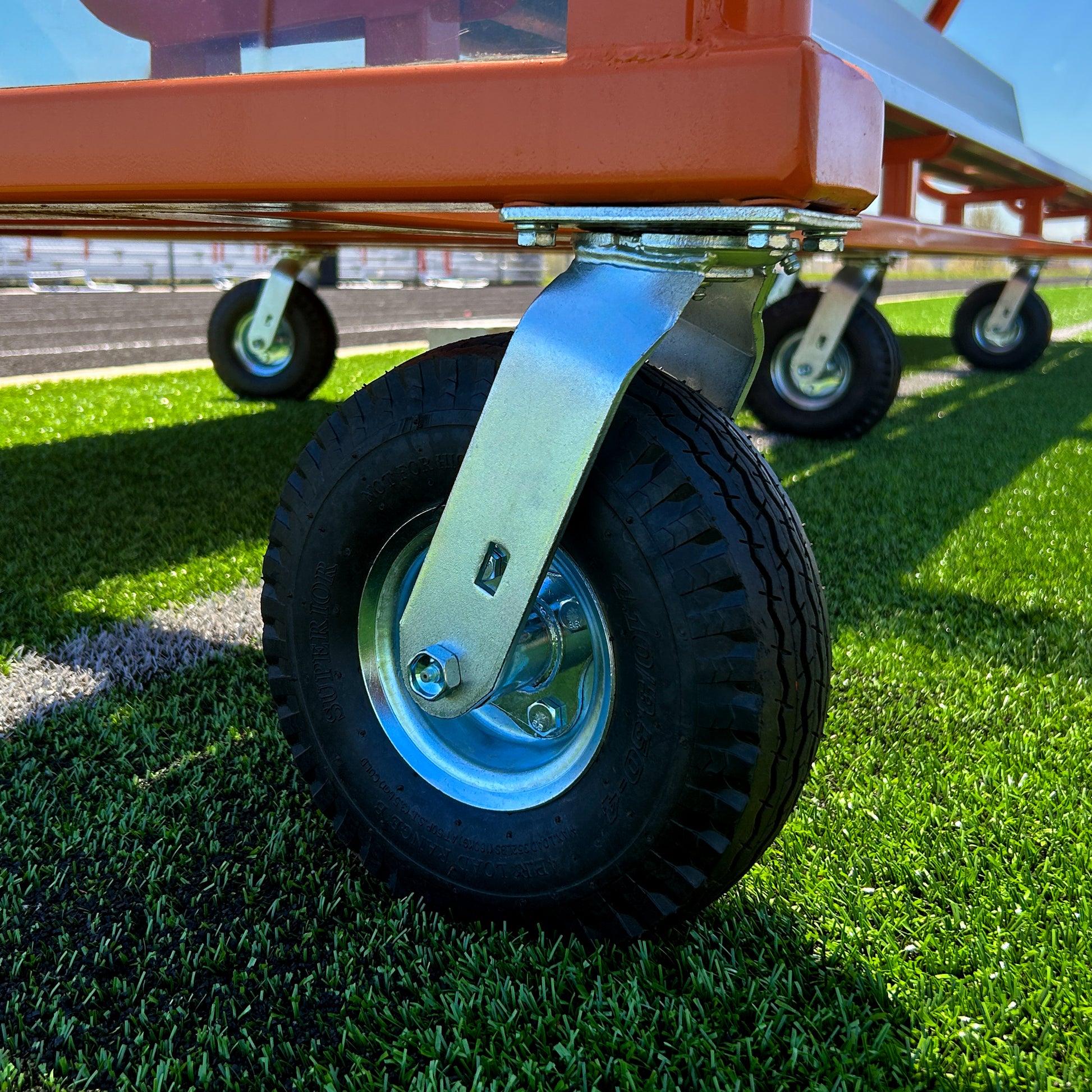 Close-up of a sturdy black rubber caster wheel affixed to the metal frame of the 16 Traditional Heavy-Duty Team Shelter cart on artificial green turf; designed for moving equipment, with additional wheels visible in the background.