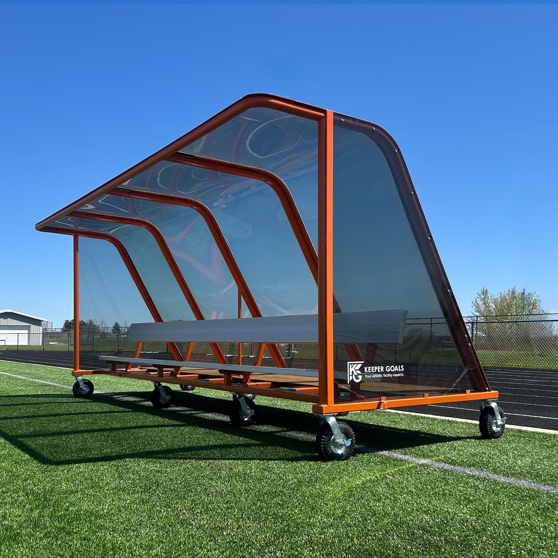 The 16 Traditional Heavy-Duty Team Shelter, featuring a modern orange frame and transparent angled canopy, sits on synthetic turf under a clear blue sky. Built for durability, this portable sports bench is mounted on wheels for easy transport.