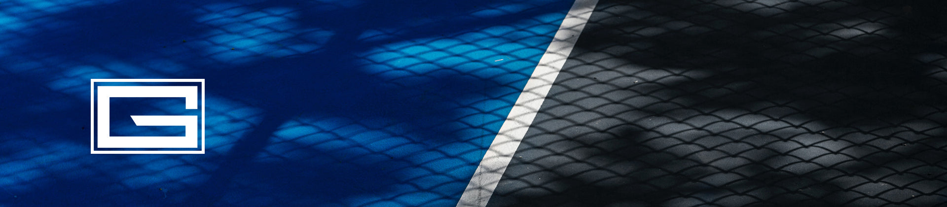 A blue and black sports court divided by a white line, with chain-link fence shadows cast across the surface. A white square logo with the letter G is on the left side of the image.