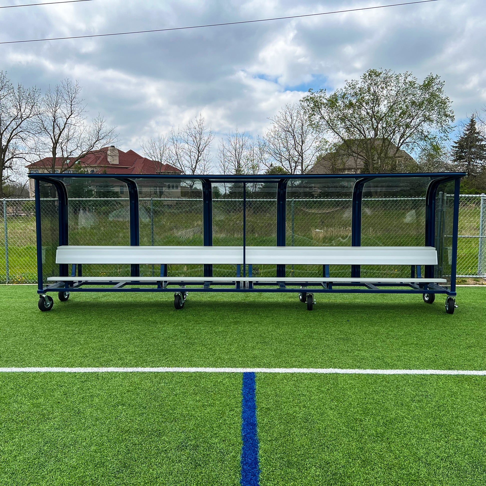 The 20 European Heavy-Duty Team Shelter, equipped with wheels, is positioned on artificial turf at a sports field, with a chain-link fence and houses in the background under a partly cloudy sky.