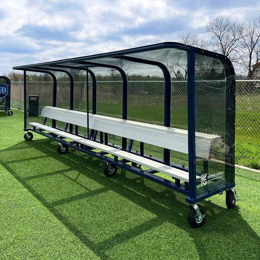The 20 European Heavy-Duty Team Shelter, featuring a metal frame, clear plastic sides, and interior seating, stands on artificial turf by a chain-link fence and trees beneath a cloudy sky.