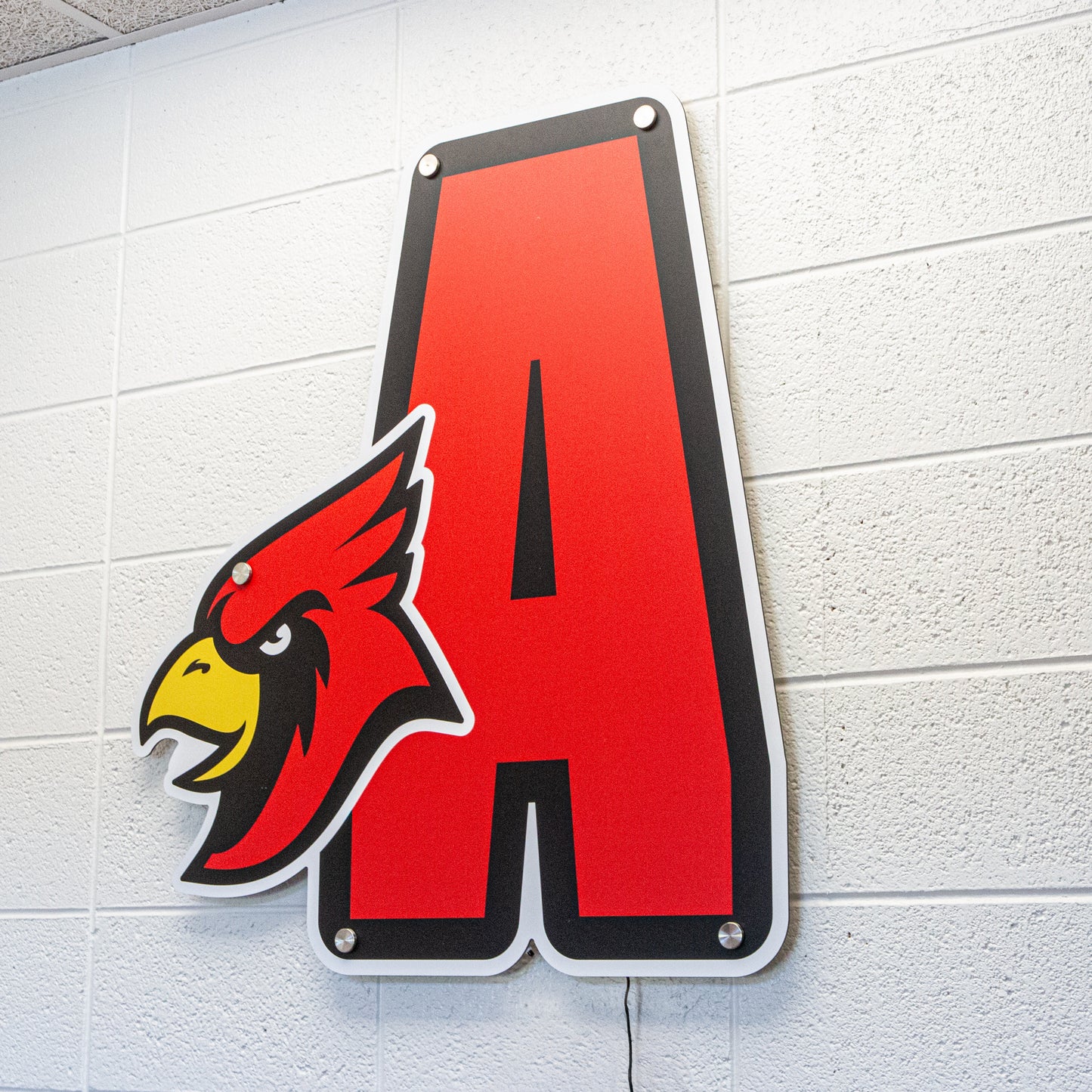 A 3D Signage or Lettering of a large red letter A is mounted on a white brick wall, featuring the head of a red cardinal bird outlined in black on the left side for enhanced visual depth.