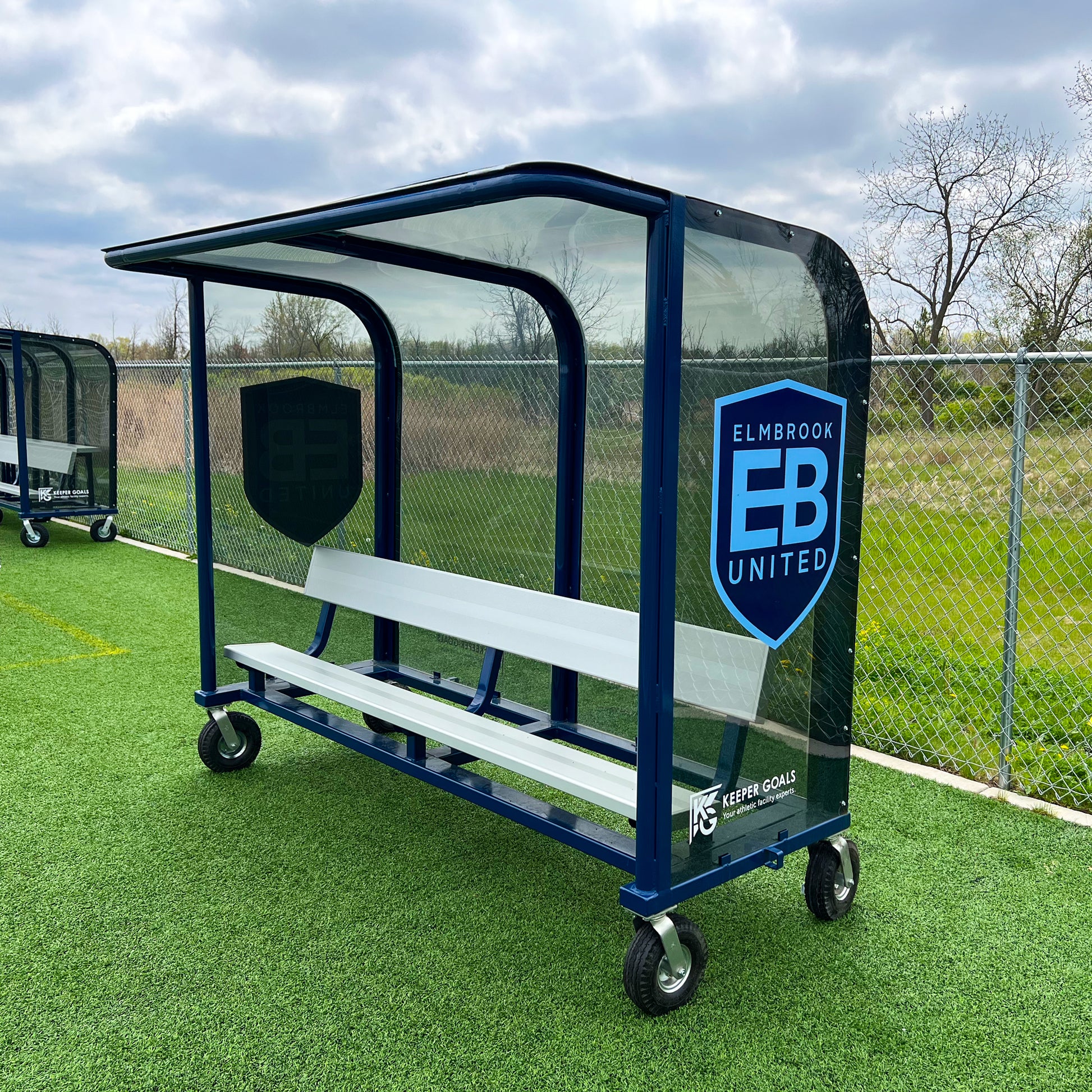 The 8 European Heavy-Duty Team Shelter in blue and white with a clear roof and side panels sits on artificial turf. With wheels, durable build, and the Elmbrook United logo, its ideal for teams needing reliable outdoor seating.