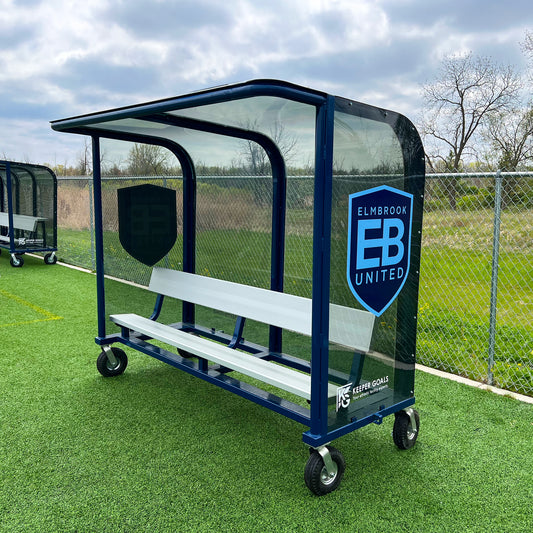 The 8 European Heavy-Duty Team Shelter in blue and white with a clear roof and side panels sits on artificial turf. With wheels, durable build, and the Elmbrook United logo, its ideal for teams needing reliable outdoor seating.