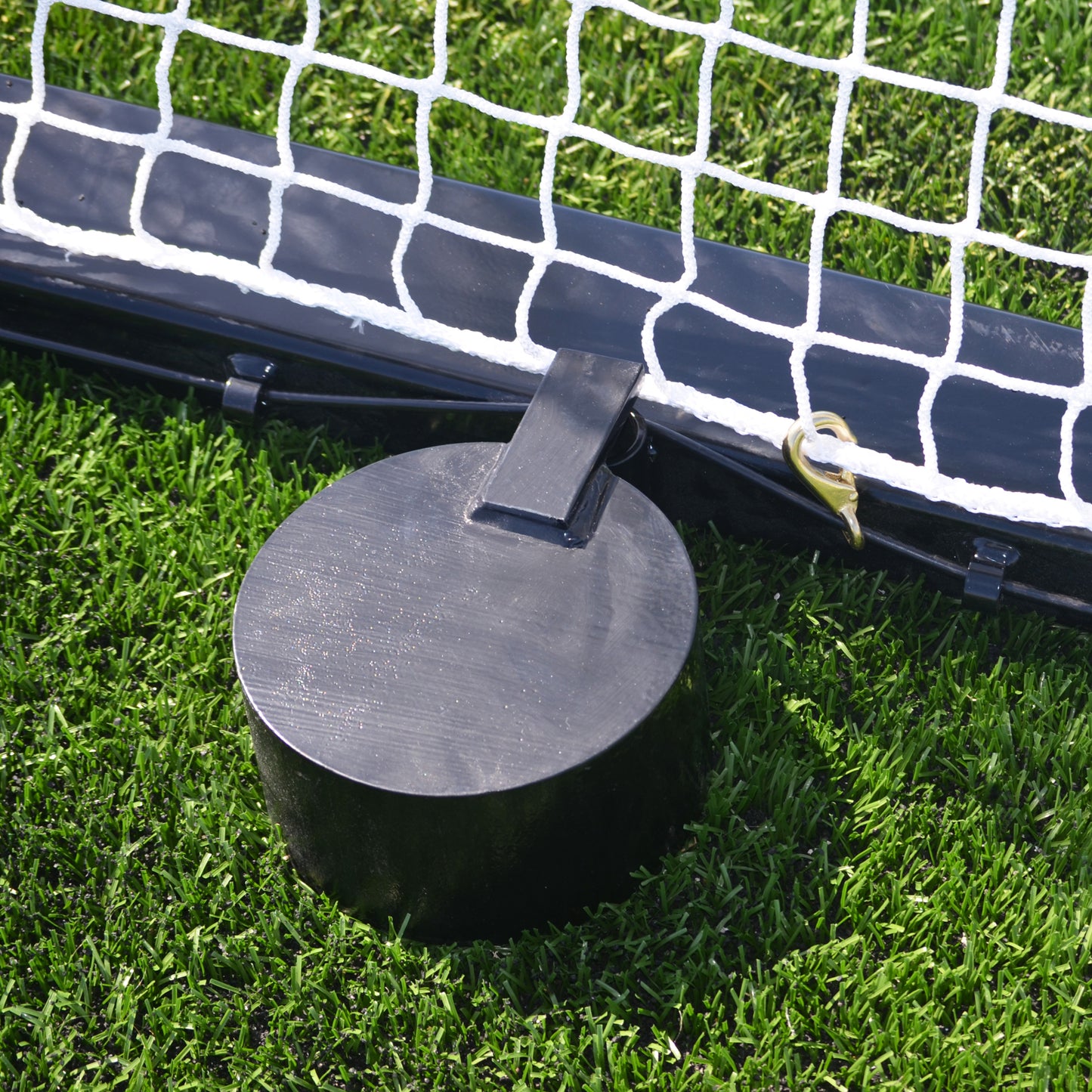 A close-up of a 28 lb. Anchor Weight securing a net to green grass, with part of the white net and black frame visible.