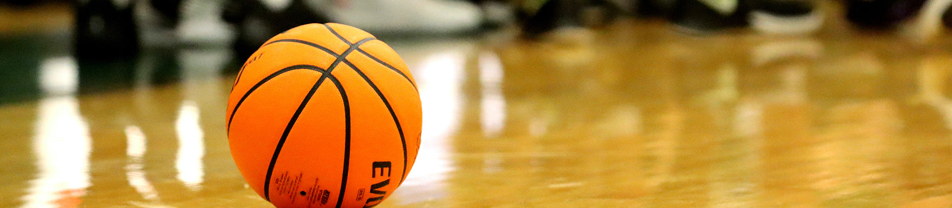 A close-up of an orange basketball resting on a shiny indoor court floor, with blurred players or spectators in the background.