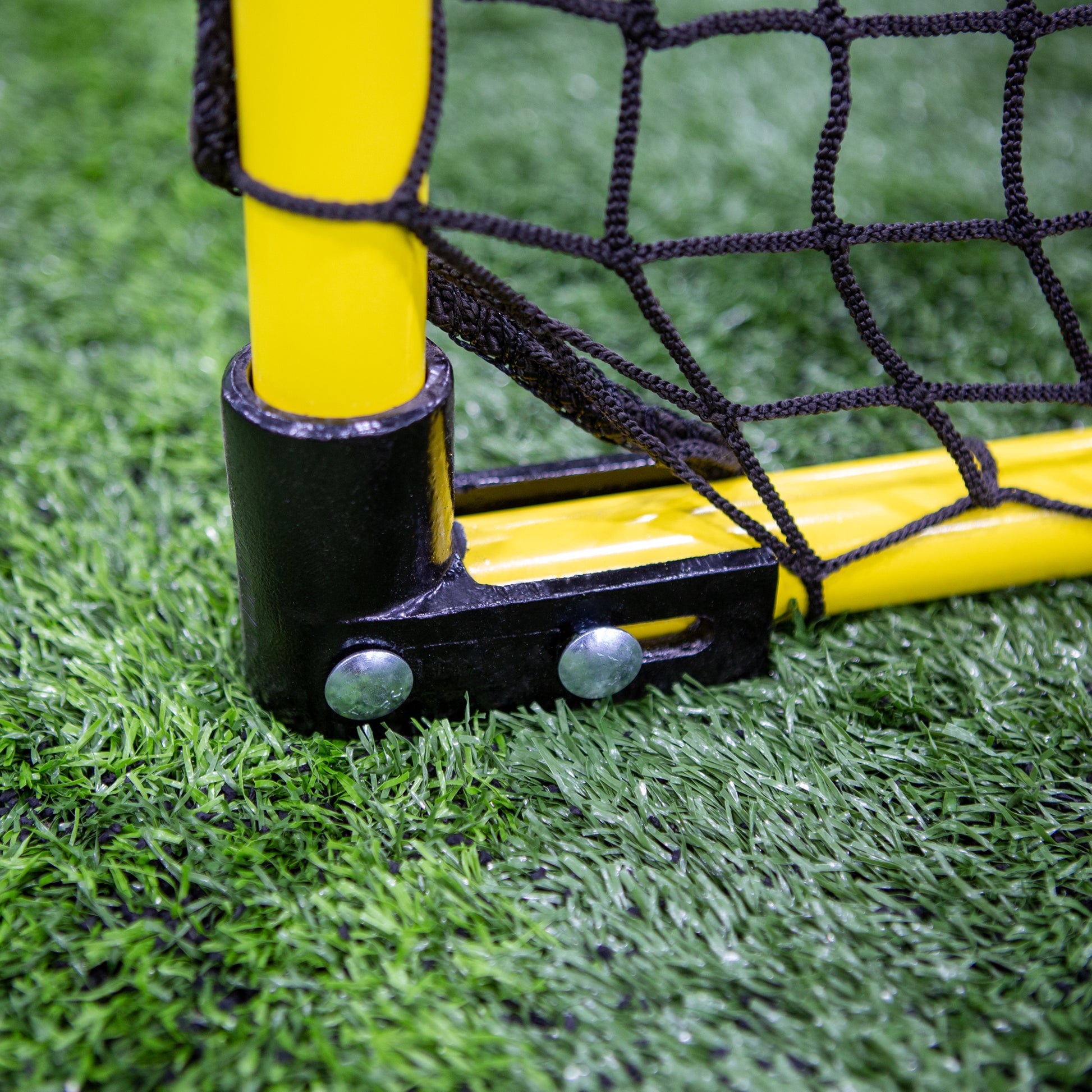 Close-up of the bottom corner of a Folding Aluminum Soccer Goal in yellow with black netting, secured by bolts on artificial green grass—perfect for those looking for portable small soccer goals.