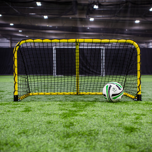 A Folding Aluminum Soccer Goal in yellow and black stands on indoor artificial turf with a white and green soccer ball against its net, while another full-size goal is visible in the background.