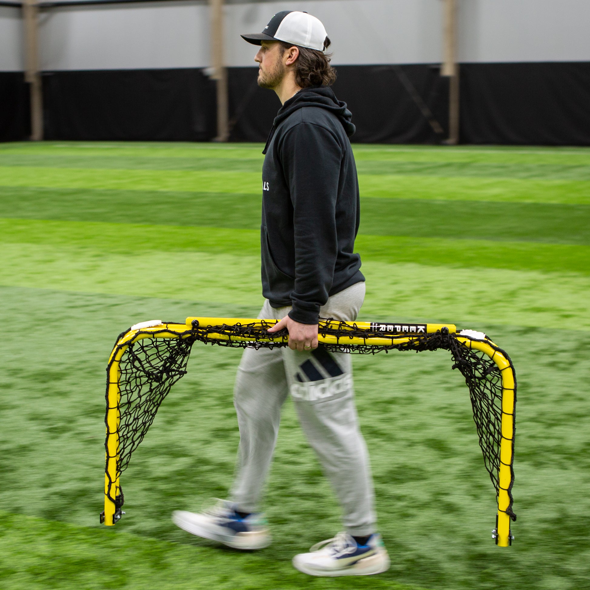 A person wearing a black hoodie, gray sweatpants, and a cap carries Folding Aluminum Soccer Goals across an indoor field with green artificial turf.