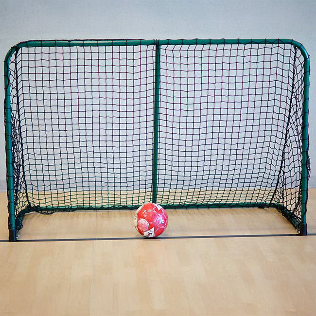 A red and white soccer ball sits in front of a green, rectangular Folding Aluminum Soccer Goal with a black net on a light wooden floor against a pale wall.