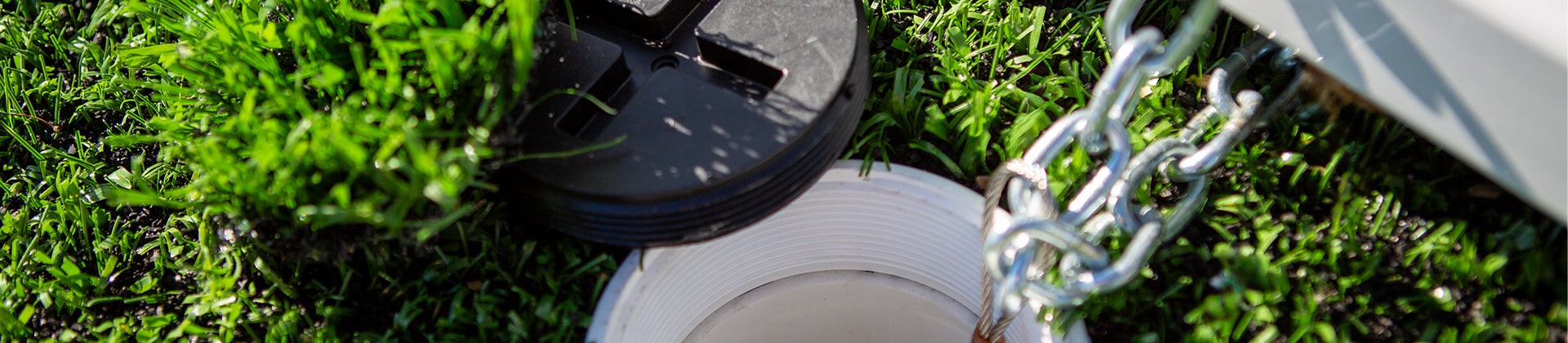 A close-up view of an RV sewer hose connected to a white ground pipe on green grass, with a black plastic cover lying nearby and a metal chain in the foreground.