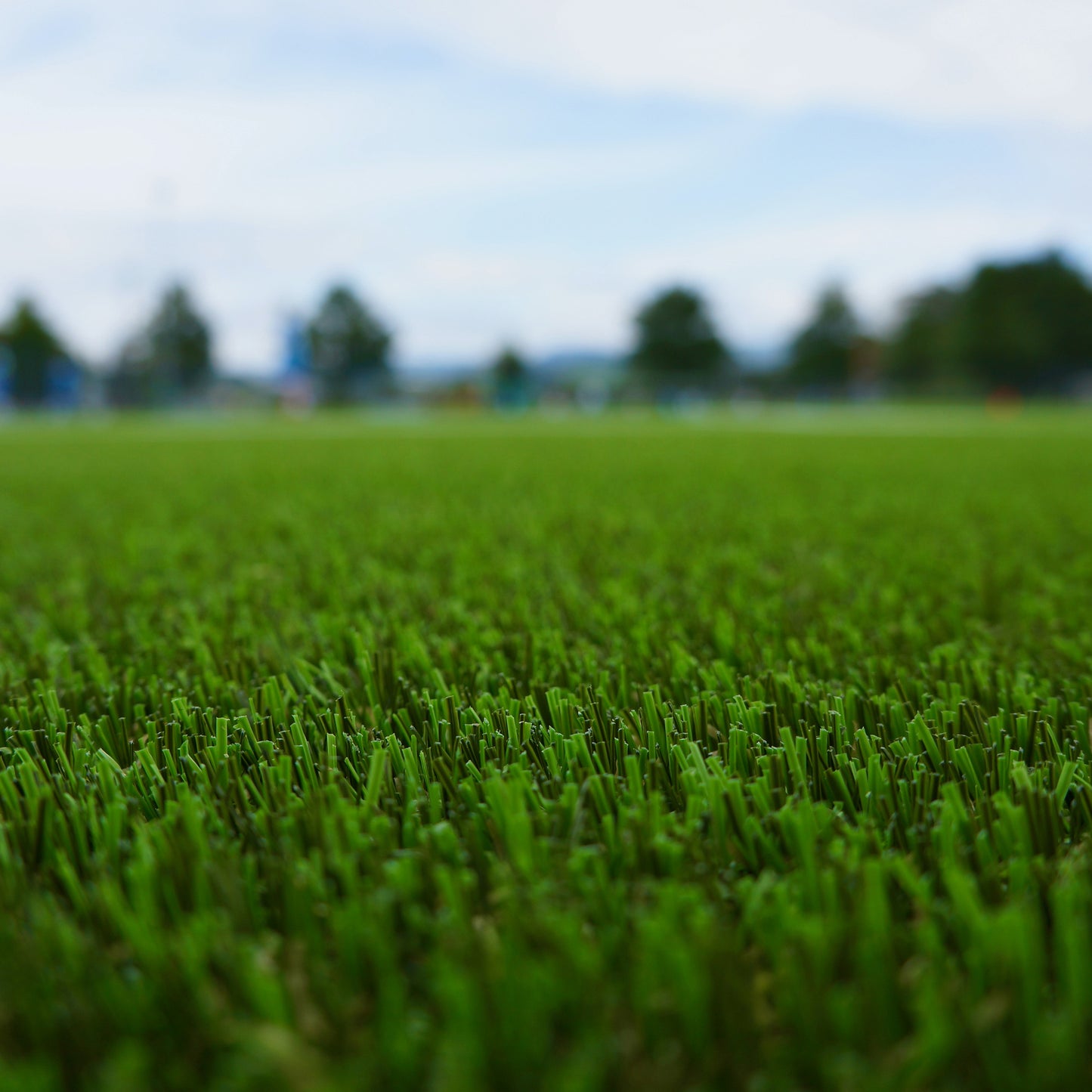 Close-up of green Artificial Turf on an outdoor sports field, with trees and a cloudy sky softly blurred in the background—delivering a realistic playing experience.