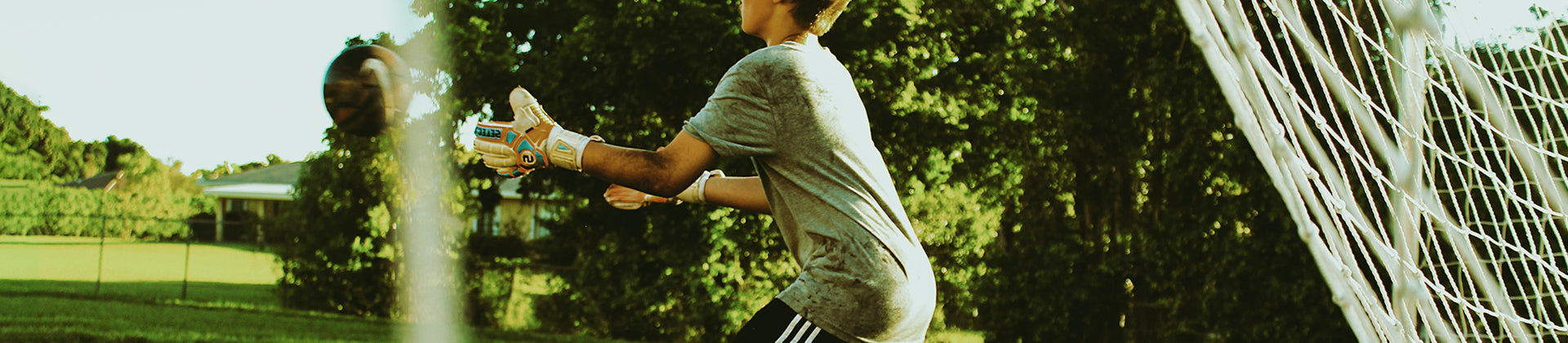 A soccer goalie in a gray shirt and gloves prepares to catch a ball while standing in front of a goal net on a grassy field with trees in the background.