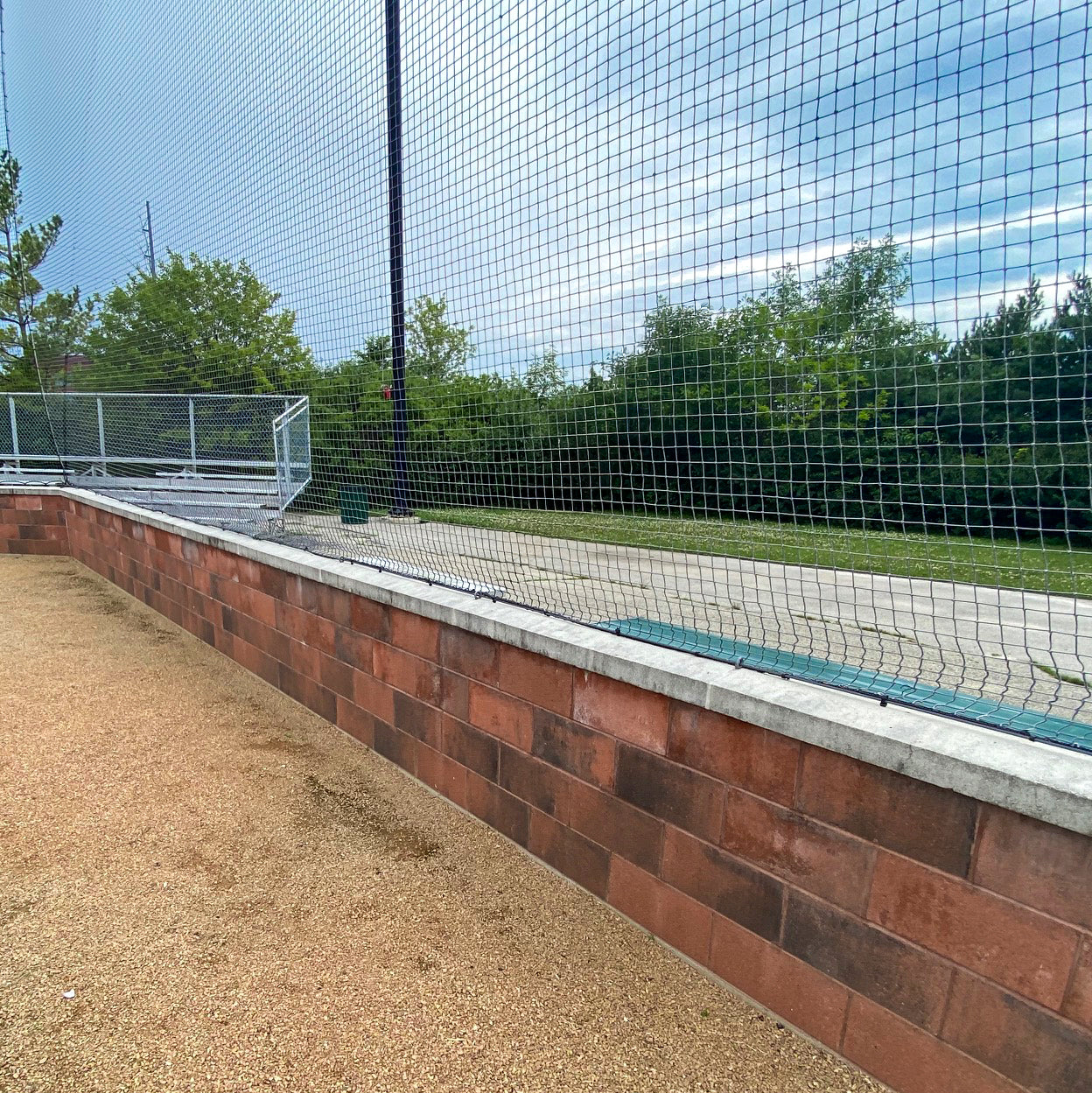 A brick wall with a concrete top borders a gravel path, topped with Custom Engineered Backstop and Backup Netting Systems; green trees and a cloudy sky create the perfect setting for any multi-sport facility.