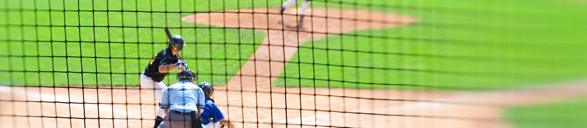 Baseball game viewed through a protective net; a batter stands at home plate ready to hit, with a catcher and umpire crouched behind him and a pitcher in the distance on the mound.