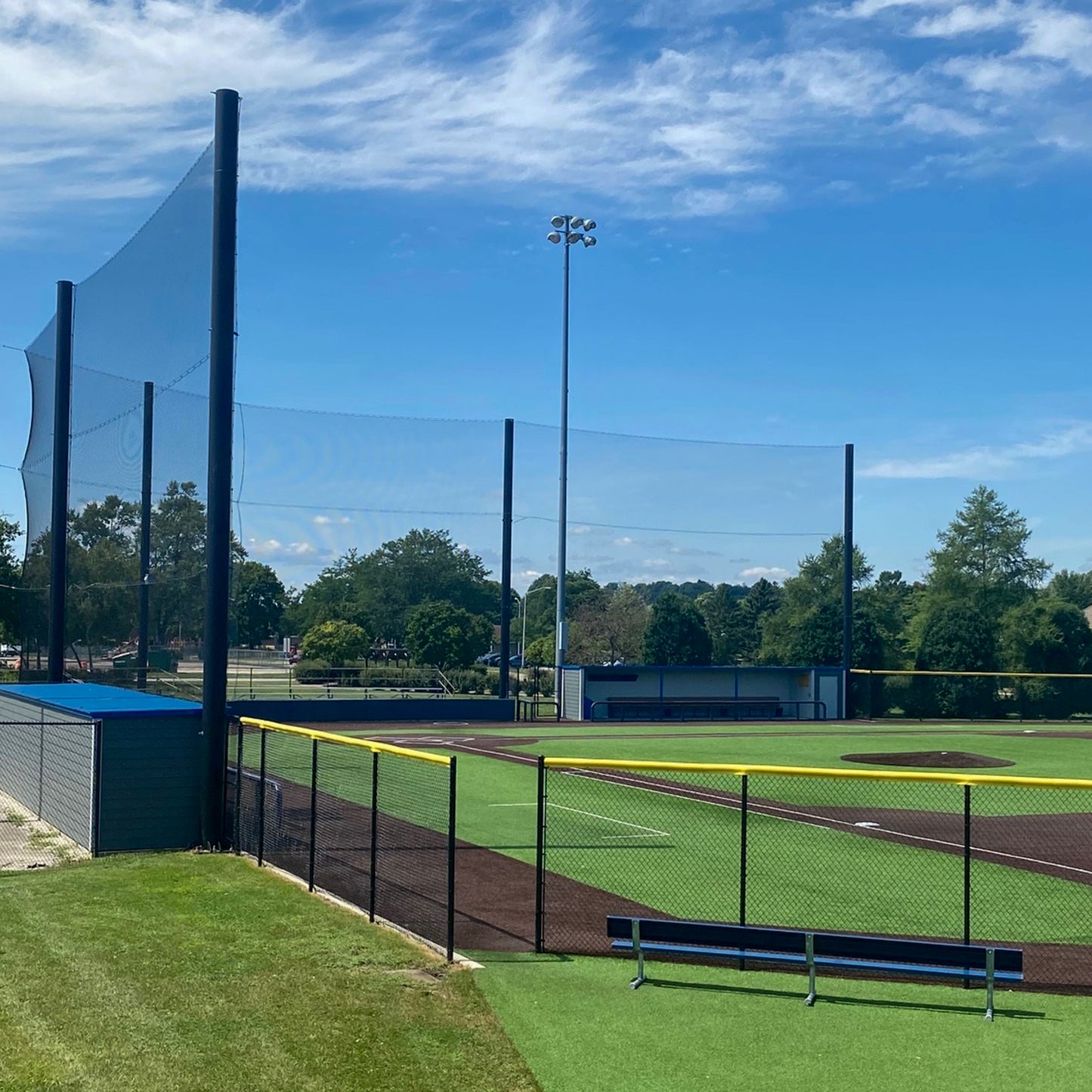 A sunny day at an empty baseball field with green artificial turf, a pitcher’s mound, and a tall Custom Engineered Backstop and Backup Netting System. A yellow-lined fence, light poles, and trees stand under a partly cloudy sky.