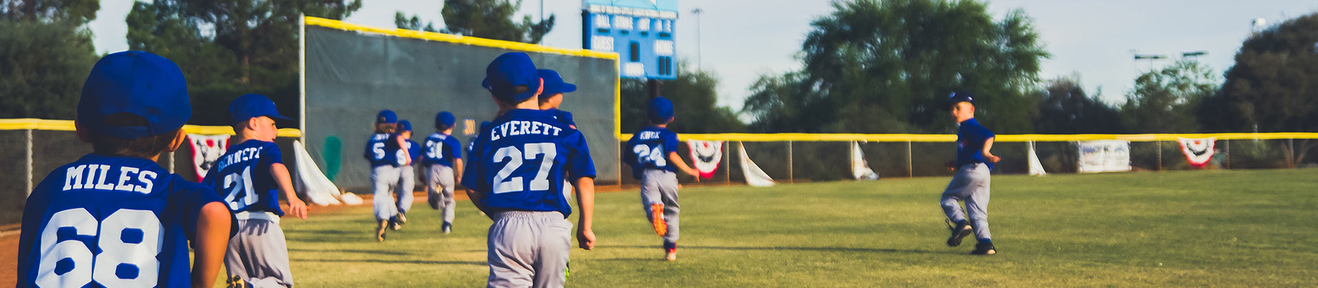 A group of young children in blue jerseys and gray pants run across a grassy baseball field, some heading toward a scoreboard and net in the background on a sunny day.