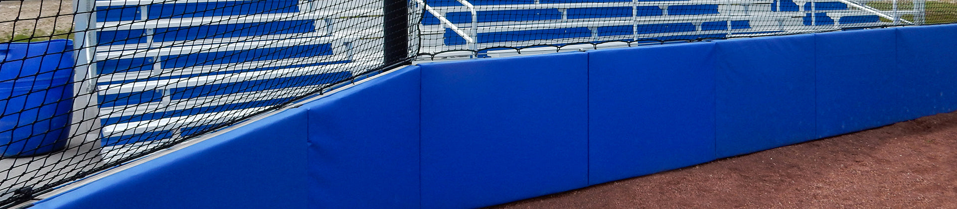 Blue padded fence and black safety net in front of white and blue metal bleachers at a sports field, with reddish dirt in the foreground.