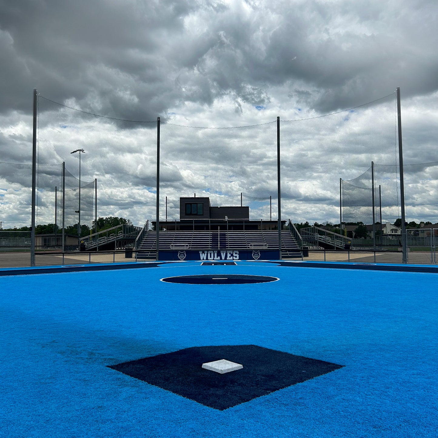 A baseball field with blue turf sits under a cloudy sky. Behind home plate, Custom Engineered Backstop and Backup Netting Systems frame empty bleachers labeled Wolves and the press box in the background.