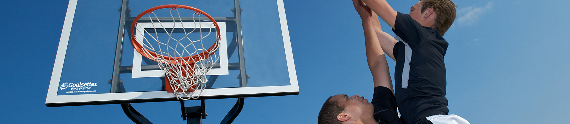Two young basketball players jump near an outdoor basketball hoop, with one attempting to block the other’s shot. The sky is clear and blue in the background.