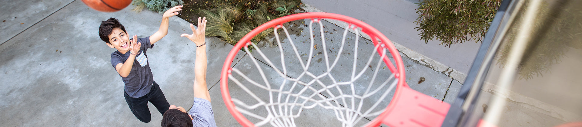 A boy jumps to shoot a basketball toward a hoop while another person tries to block him on an outdoor court, viewed from above near the rim.