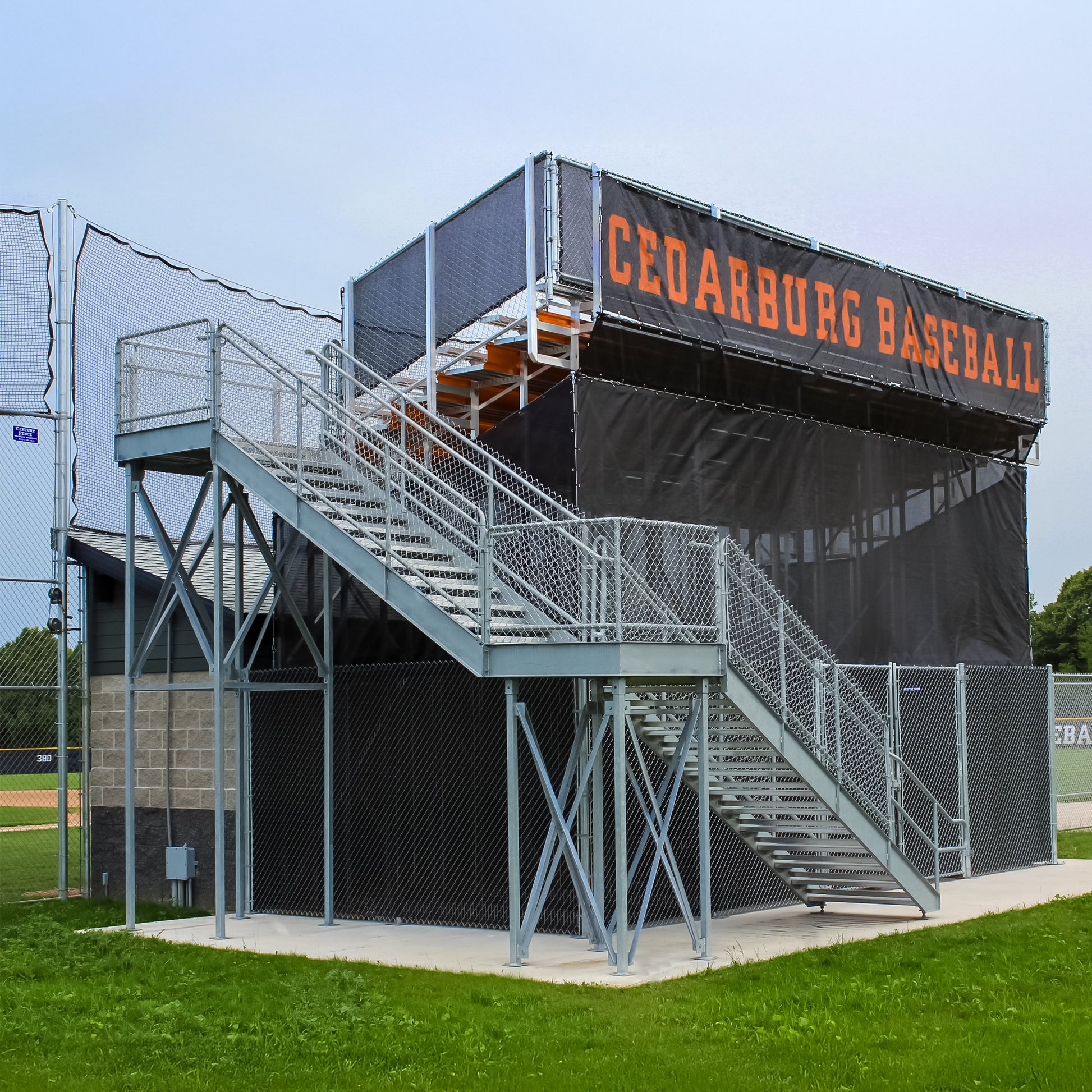 A Bleacher Wrap installed on a metal bleacher with stairs at a baseball field displays Cedarburg Baseball in black, features custom stadium graphics, and is enclosed by chain-link fencing.