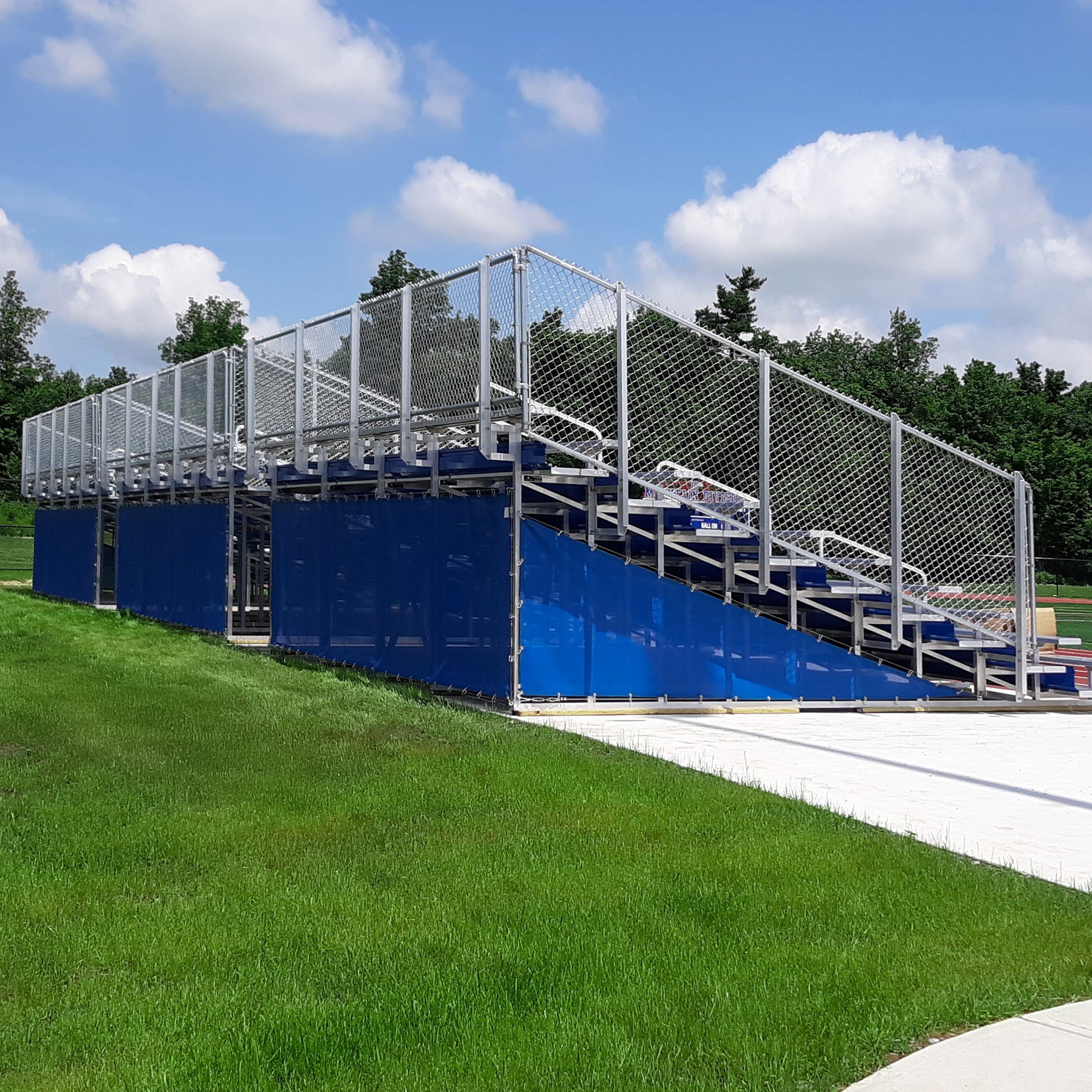 Bleachers featuring blue paneling and a custom Bleacher Wrap are placed on grass by a sidewalk, with trees and a partly cloudy blue sky in the background.