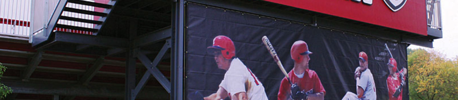 A large outdoor banner displays five baseball players in action poses, attached to a metal structure beneath a red stadium section.