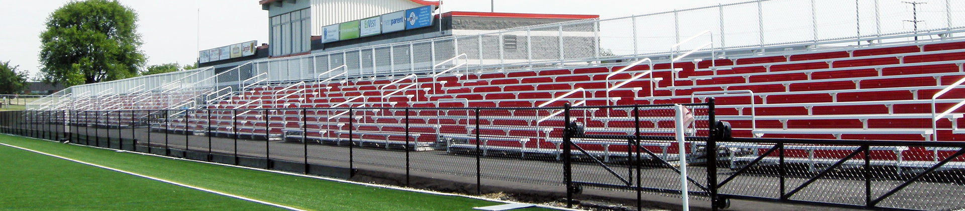 Long, empty red and white bleachers behind a black chain-link fence at a sports field, with a building and trees in the background under a clear sky.