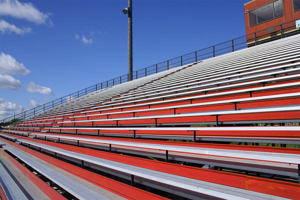 Empty red and white stadium bleachers under a bright blue sky with some clouds, a tall light pole, and a press box visible in the background.