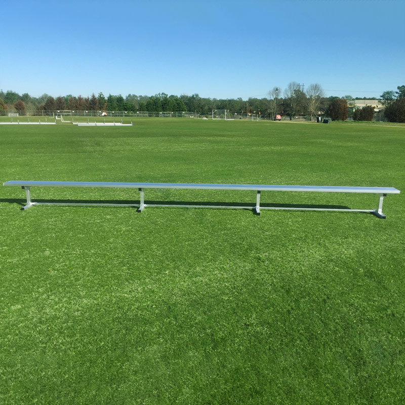 A Budget Portable Aluminum Team Bench with no backrest sits on green grass under a clear blue sky, with trees and goalposts in the background.