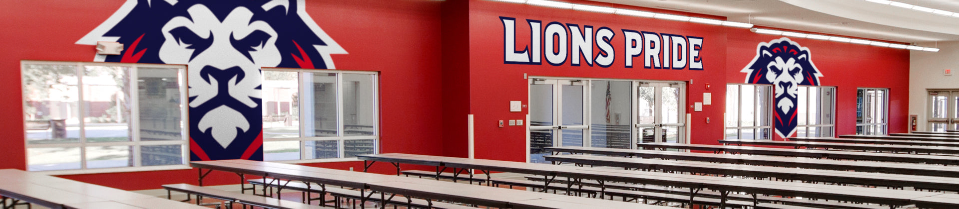 A school cafeteria with long rows of empty tables and benches, red walls featuring a large lion mascot graphic and bold text reading LIONS PRIDE above windows.