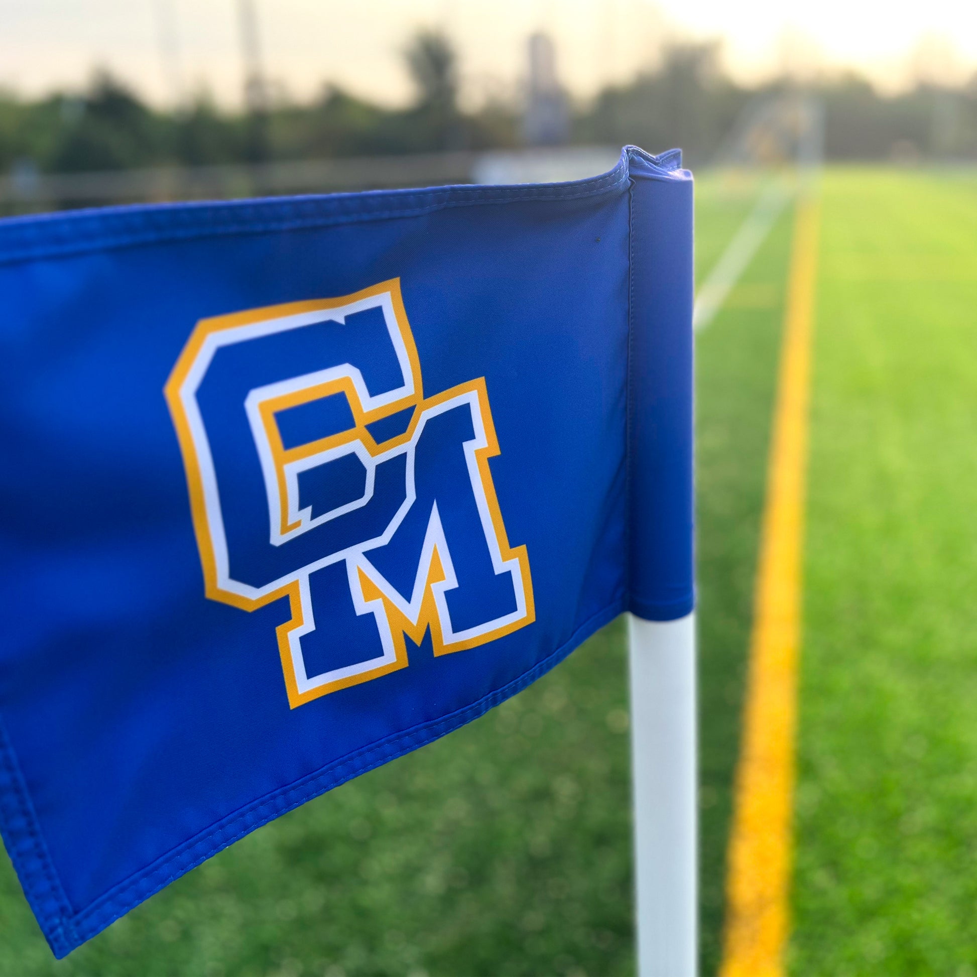A Custom Soccer Corner Flag in blue with bold yellow and white "CM" initials stands on a green field, highlighted by sharp white and yellow lines beneath soft sunlight, showcasing premium branded soccer field equipment.