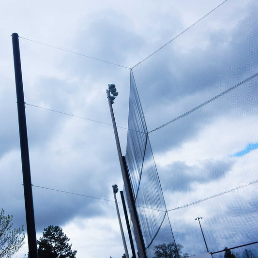 Tall black poles support a Custom Engineered Backstop and Backup Netting System, with stadium lights in the background, cloudy skies above, and trees at the lower left of this multi-sport facility.