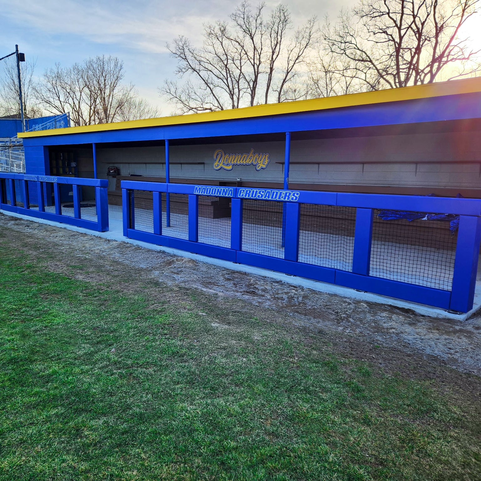A Custom Outdoor Protective Padding For Poles, Posts and Rails in bright blue and yellow with Danuboy and Middletown Crusaders on its vinyl cover, set by a grassy field and leafless trees under a clear sky.
