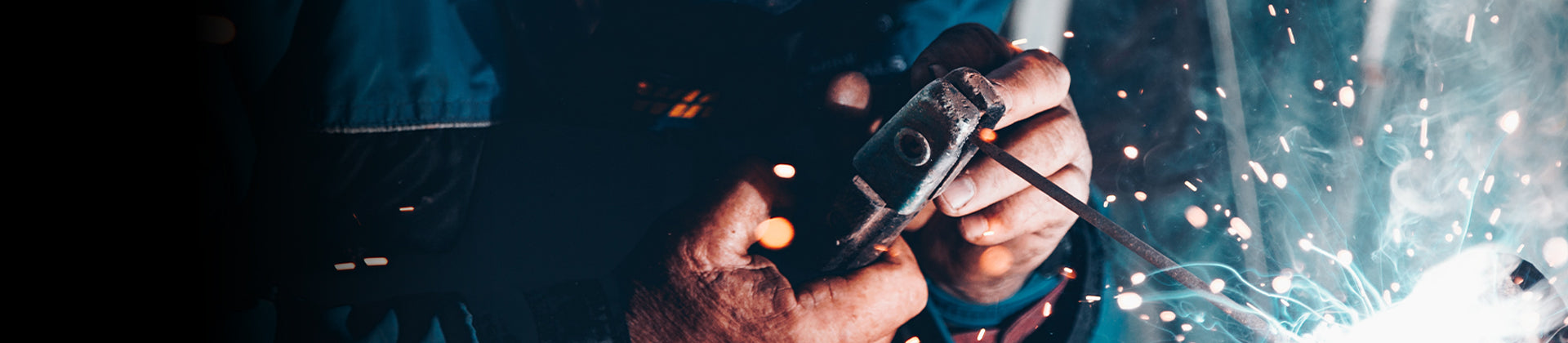 Close-up of hands holding metal parts and a tool, with bright sparks flying and blue light from welding in the background, suggesting industrial work or metal fabrication.