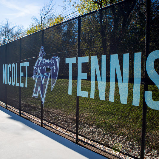 At an athletic facility, a Custom Fit Windscreen displays large “Nicolet Tennis” text and a knight mascot, providing wind blockage with trees and grass visible behind it.