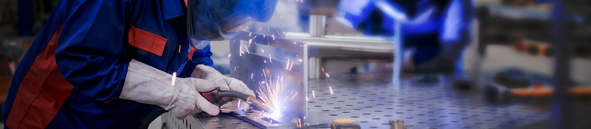 A person wearing protective gear welds metal on a workbench, producing bright sparks in an industrial workshop setting.