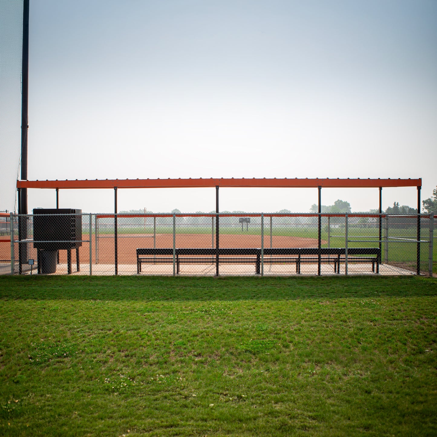 The World Series Dugout features benches behind a chain-link fence and a prefabricated steel roof for shade, facing a green field and dirt infield under clear skies.