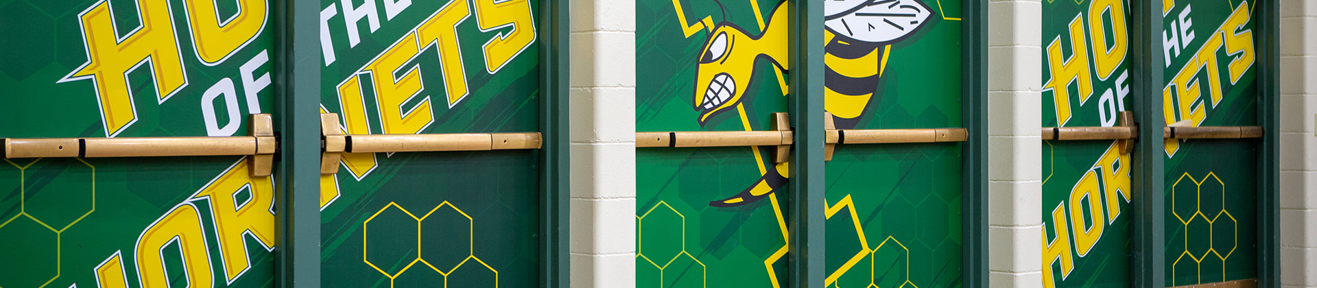 Green gym doors with the words “Home of the Hornets” and an illustrated hornet logo in yellow and black. Hexagon patterns decorate the doors, which are separated by beige columns.