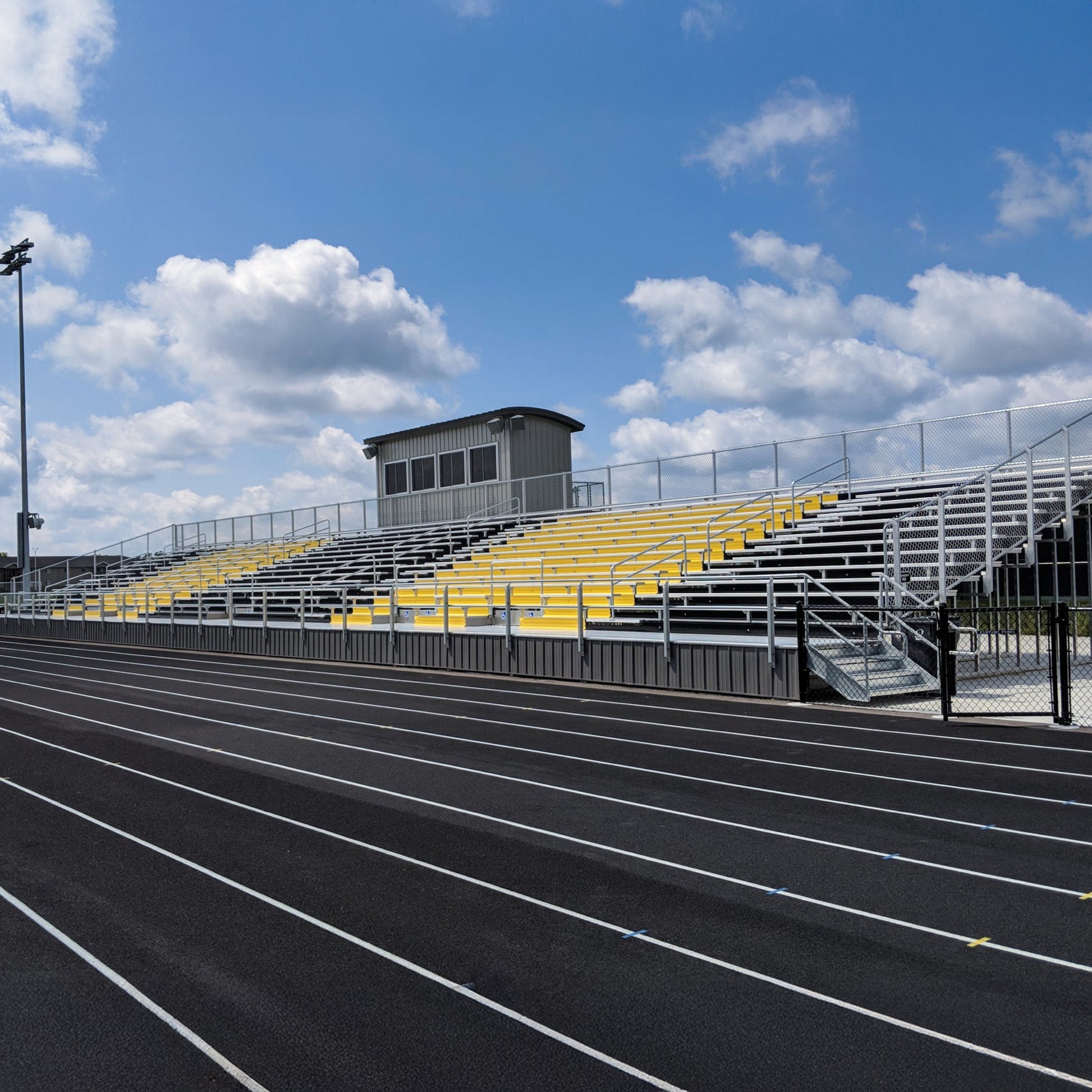 Elevated Aluminum Bleachers with yellow seating sections and robust construction overlook a multi-lane running track under partly cloudy skies. A small press box is positioned at the top of the bleachers.