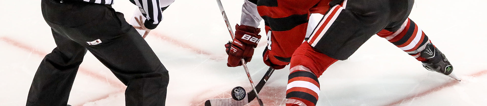 Close-up of a hockey referee dropping the puck for a face-off between two players in red and black uniforms on an ice rink.