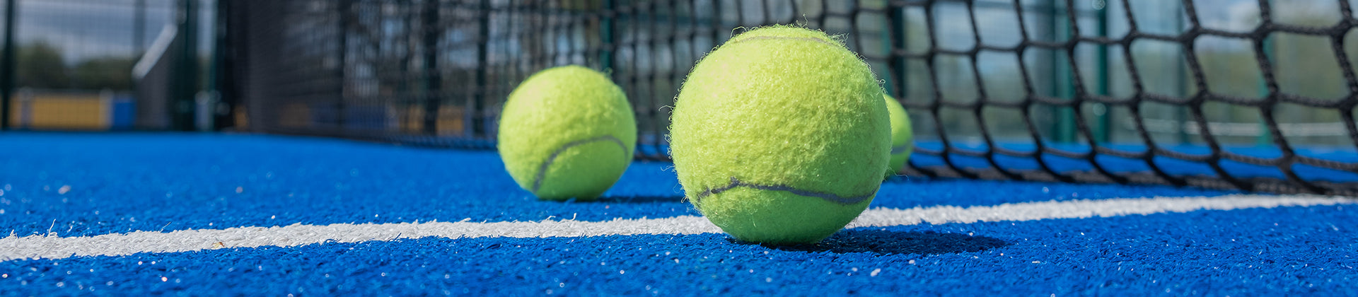 Close-up of three yellow tennis balls on a bright blue tennis court, with a tennis net in the background. The balls are scattered near the white boundary line.