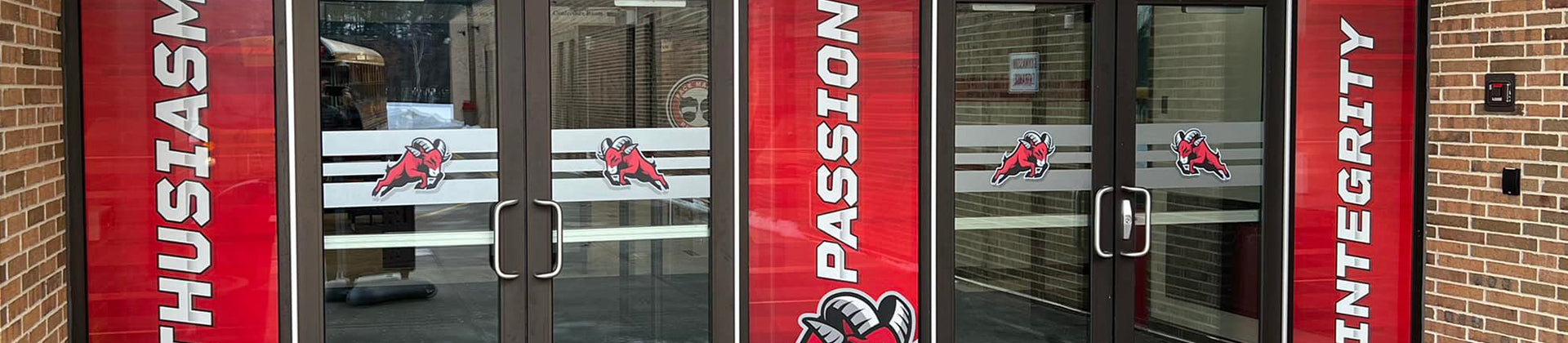 Glass double doors at a building entrance, decorated with bold red banners reading “ENTHUSIASM,” “PASSION,” and “INTEGRITY.” A mascot logo featuring a knight is displayed on the glass panels.
