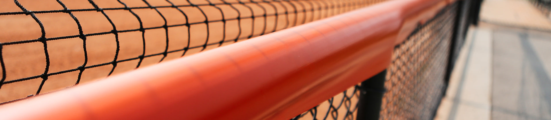 Close-up of an orange handrail along a black netted fence at a sporting field, with blurred sandy ground and pavement visible in the background.
