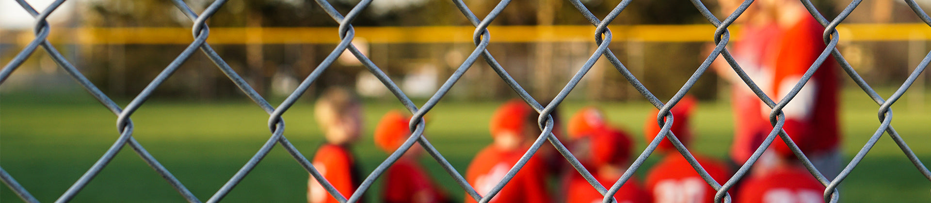A chain-link fence in sharp focus with a blurred background of children in red uniforms and hats gathered on a grassy field, possibly for a sports practice or game.
