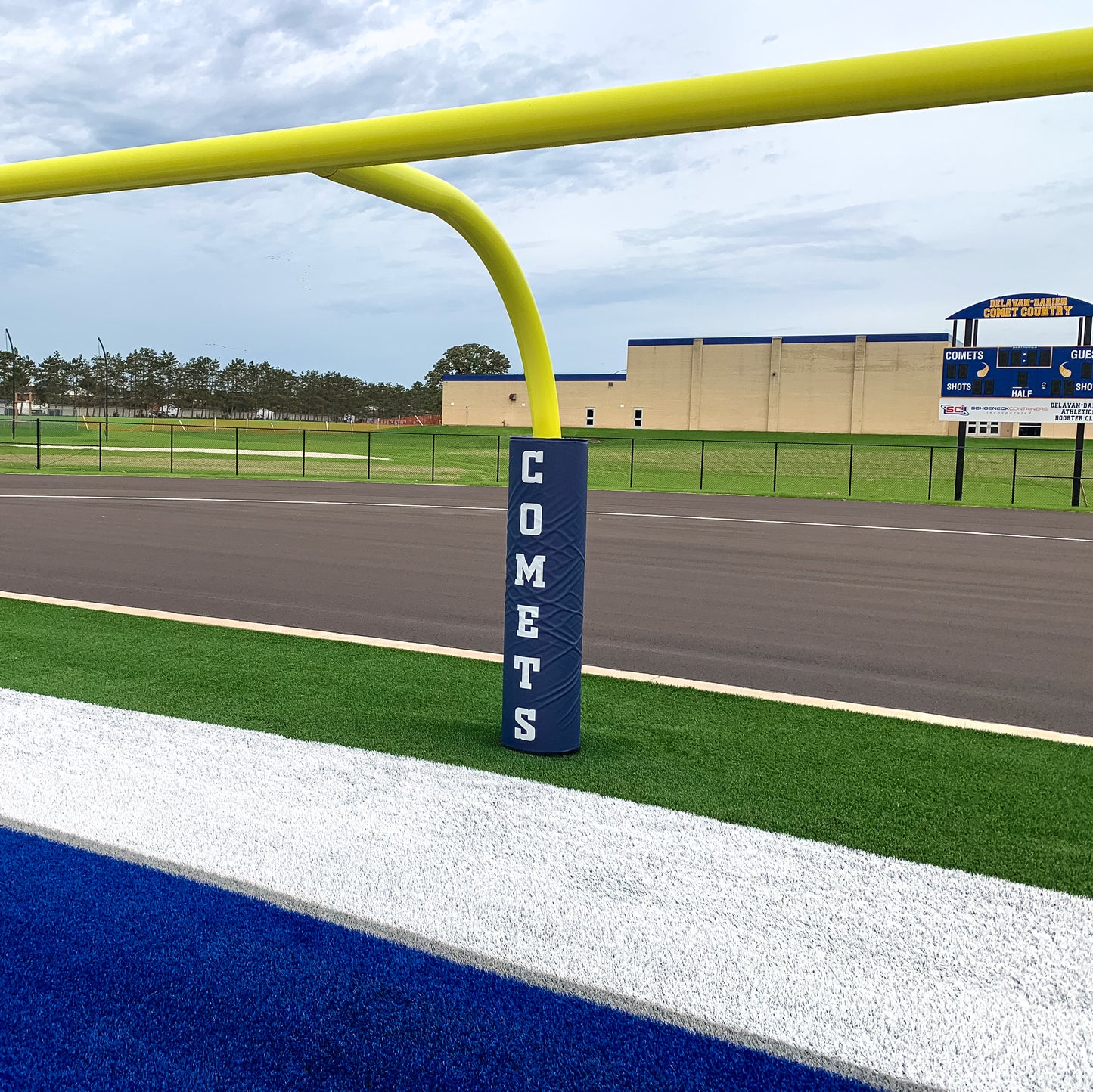 Football Goal Post Pads in blue, labeled COMETS, wrap a yellow goalpost on a green field near a blue and white end zone; the pads protect players. A scoreboard and school building appear in the background.