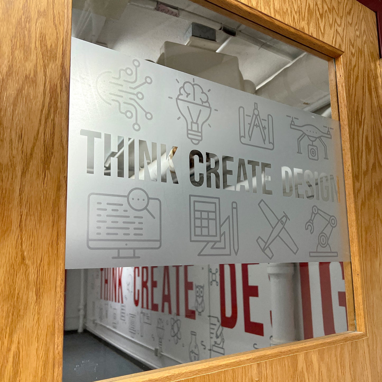A glass window on a wooden door is covered in Frosted Crystal (Etched) Vinyl with icons and “THINK CREATE DESIGN.” The modern, creative workspace inside features matching wall art and text on display.