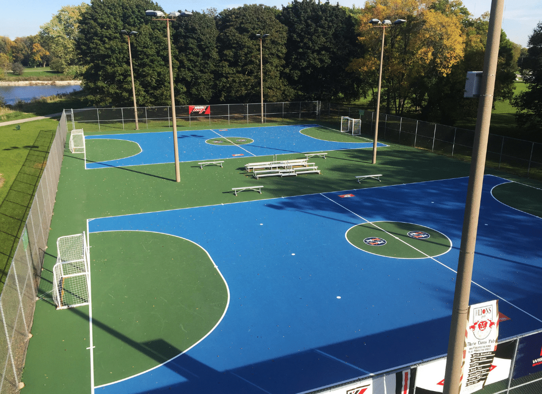 A fenced outdoor sports court with blue and green surfaces, two small soccer goals, multiple benches, and tall light poles. Trees and a body of water are visible in the background.